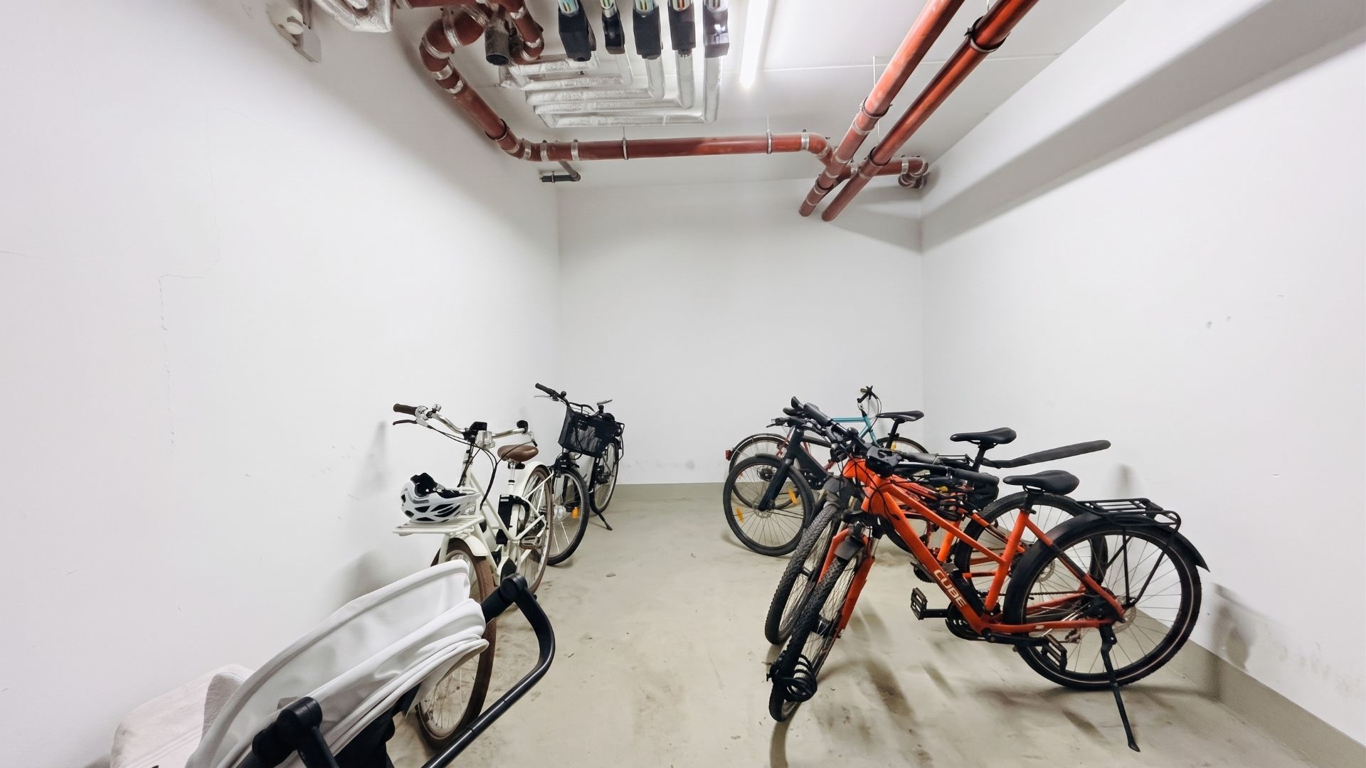 Fahrradraum mit mehreren abgestellten Fahrrädern und sichtbaren Rohrleitungen an der Decke.