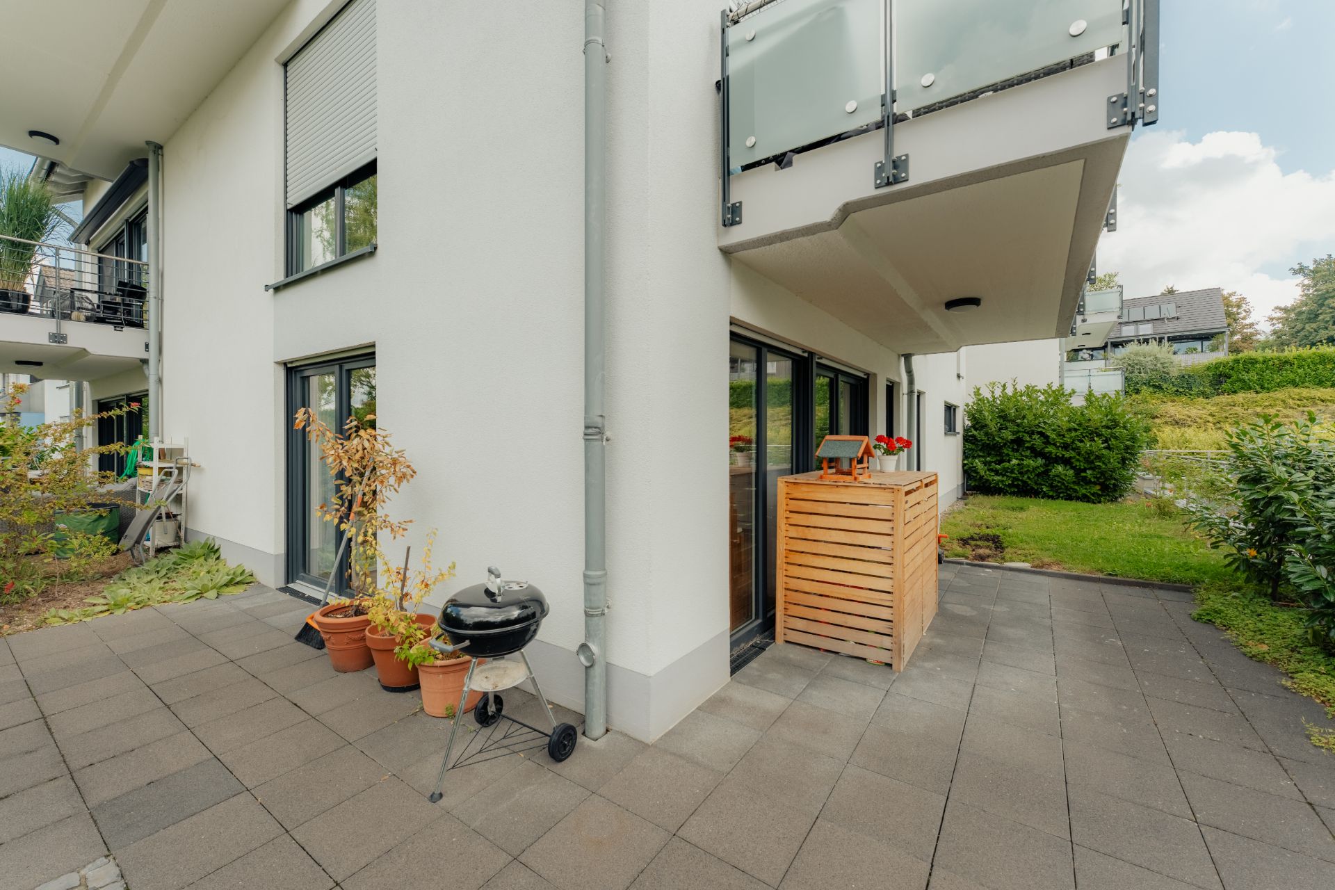 Moderne Terrasse mit Grill, Blumentöpfen und Holzverkleidung an einem weißen Wohnhaus.