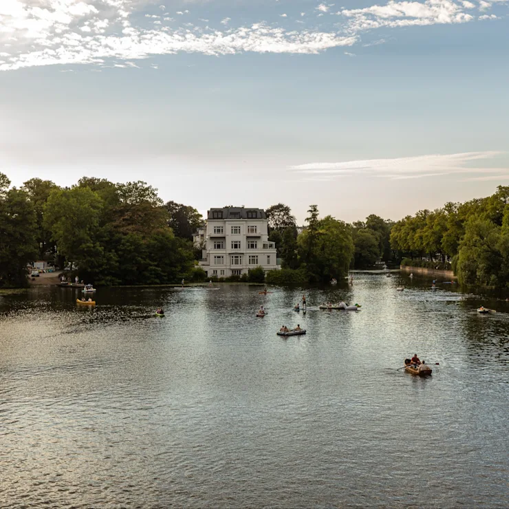 Großes Wohnhaus am See, umgeben von Bäumen, Boote und Menschen auf dem Wasser.