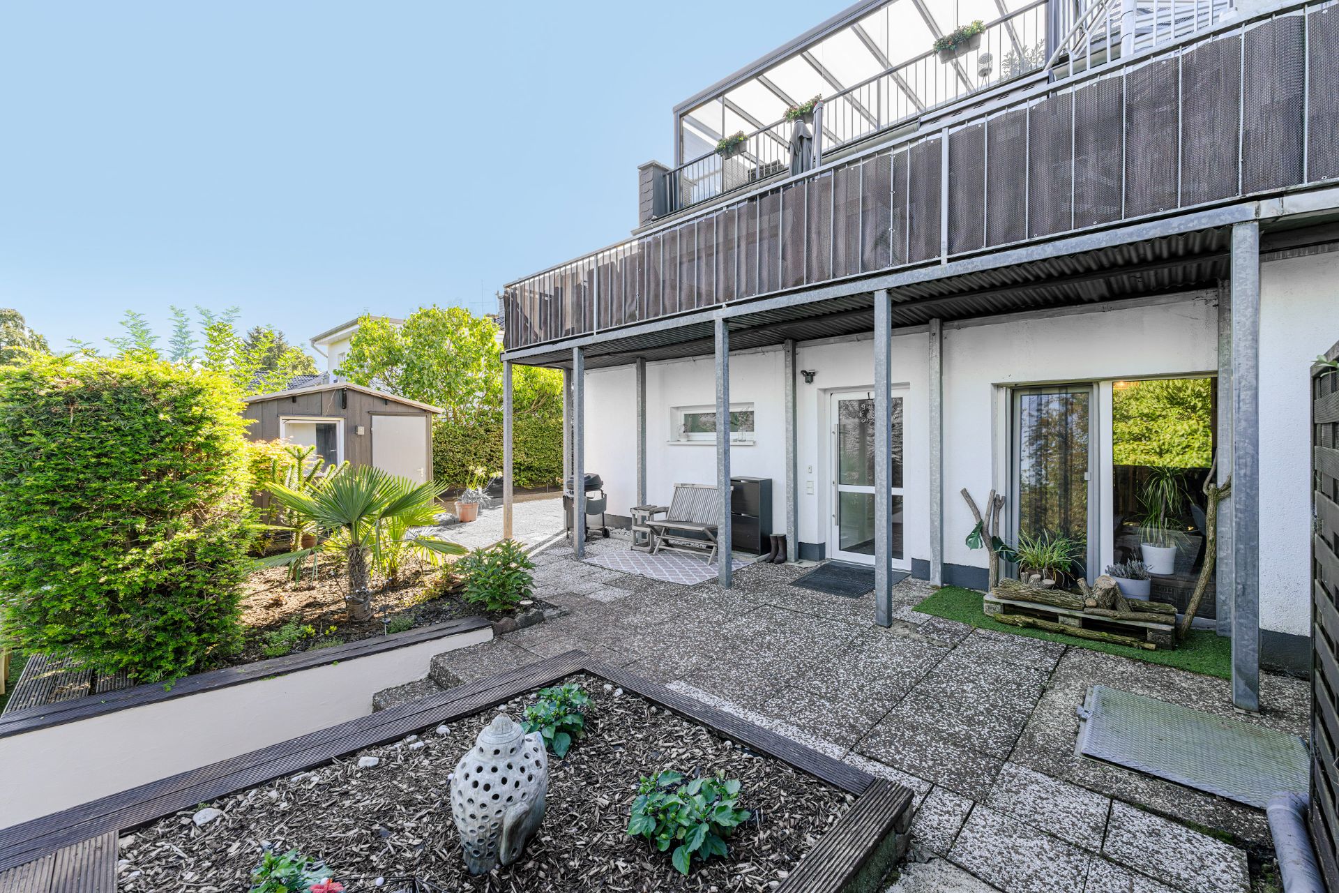 Gepflegter Gartenbereich mit Terrasse, Hochbeet, kleiner Hütte und überdachtem Balkon an einem Wohnhaus.