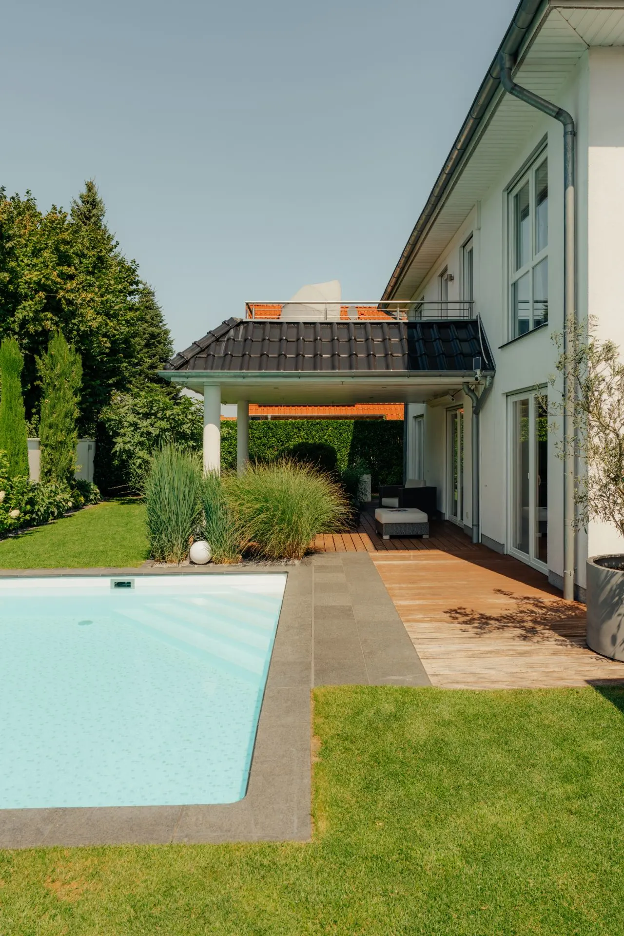 Moderner Garten mit Pool, Holzterrasse, Sitzbereich und viel Grün neben einem Wohnhaus.
