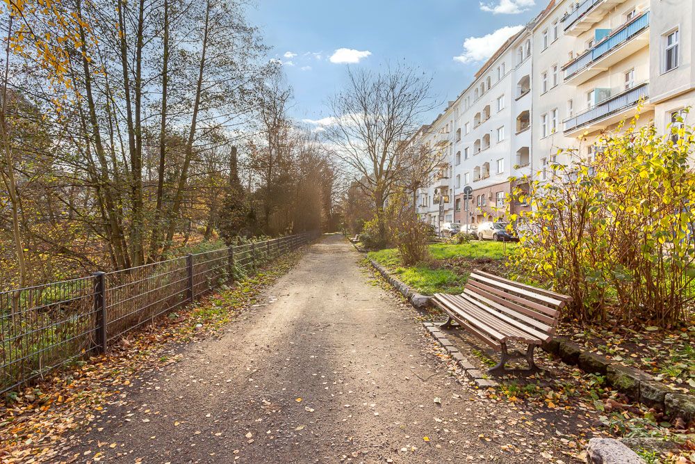Gepflasterter Weg mit Bank, Bäumen und Wohnhäusern im Hintergrund bei sonnigem Wetter.