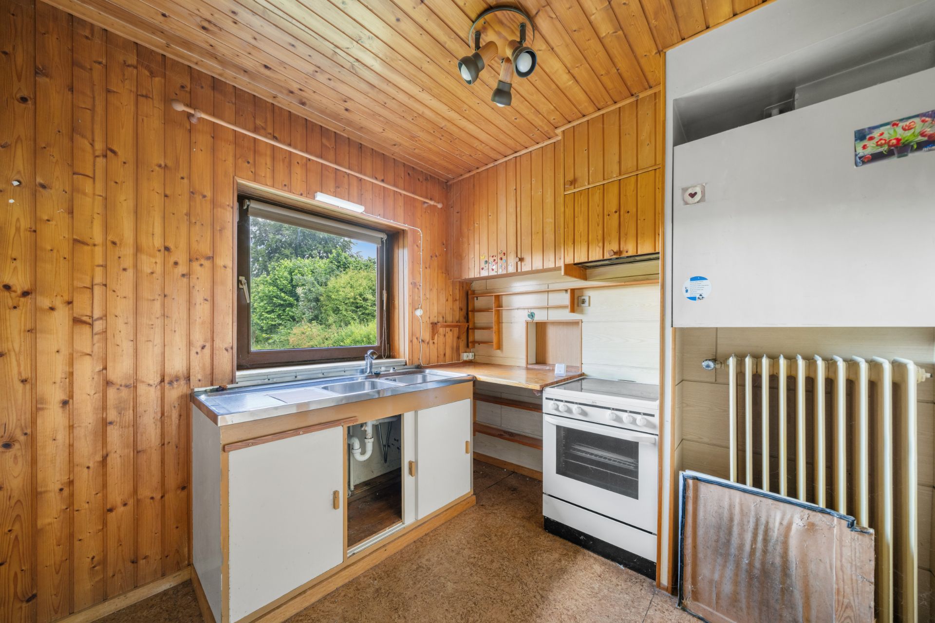 Kleine Küche mit Holzverkleidung, Fenster, Spüle, Herd und Heizkörper.