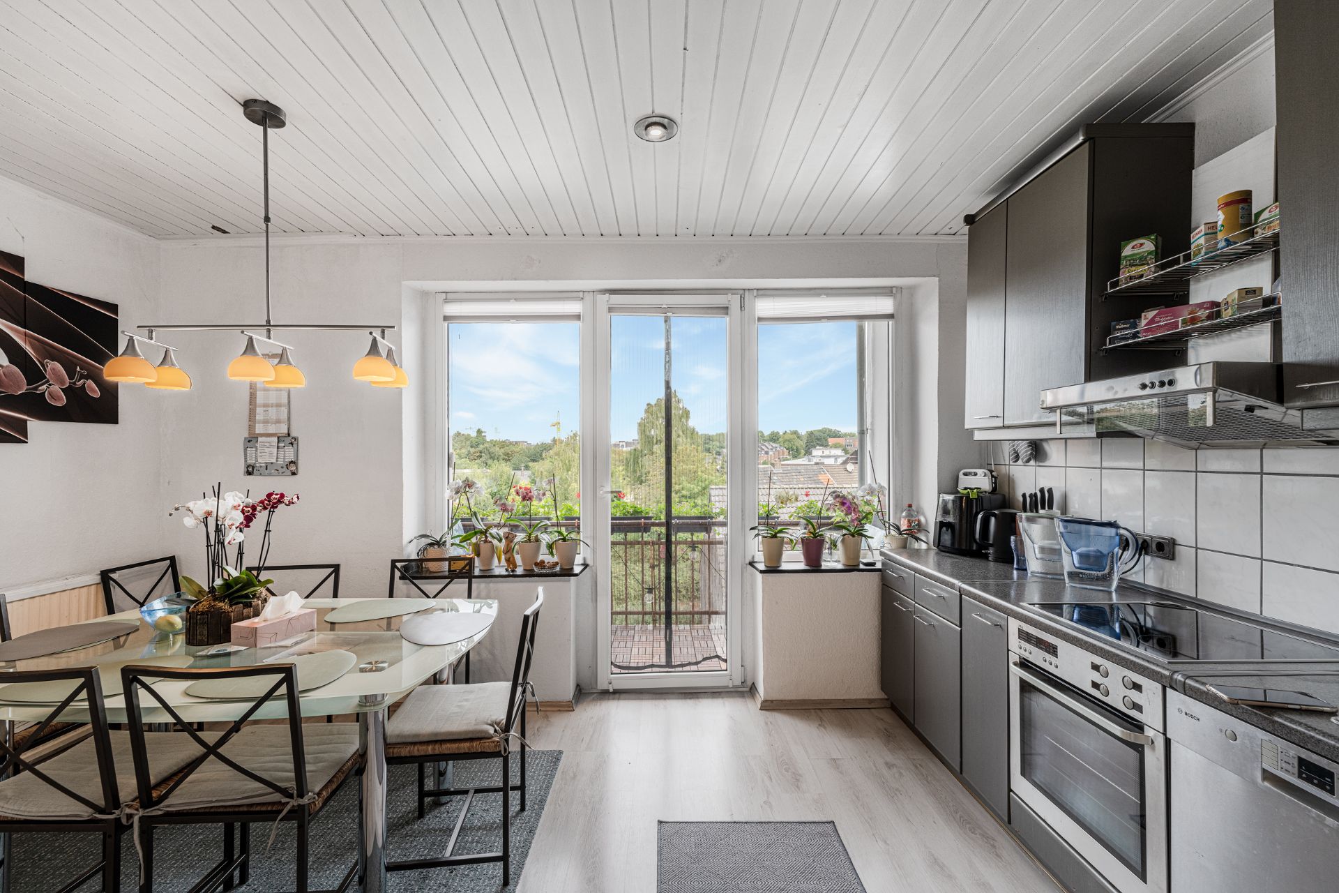 Moderne Küche mit Esstisch, großen Fenstern, Pflanzen auf der Fensterbank und Blick ins Grüne.