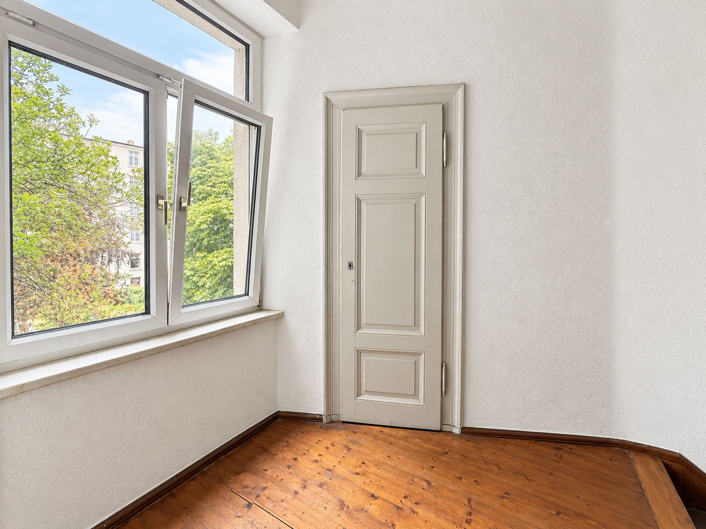 Kleiner Raum mit Holzboden, offenem Fenster und weißer Tür.