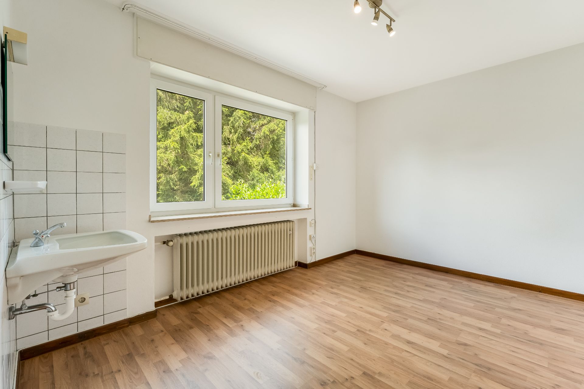 Heller Raum mit Holzfußboden, großem Fenster, Heizkörper und Wandwaschbecken.
