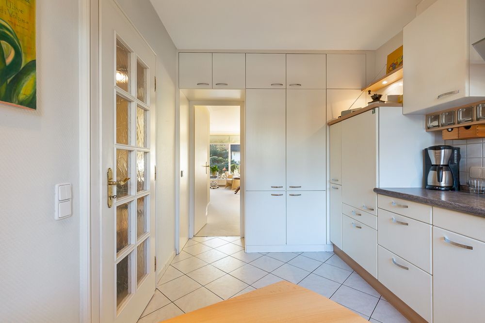 Helle Küche mit weißen Einbauschränken, Fliesenboden und einer Glastür, Blick ins Wohnzimmer.