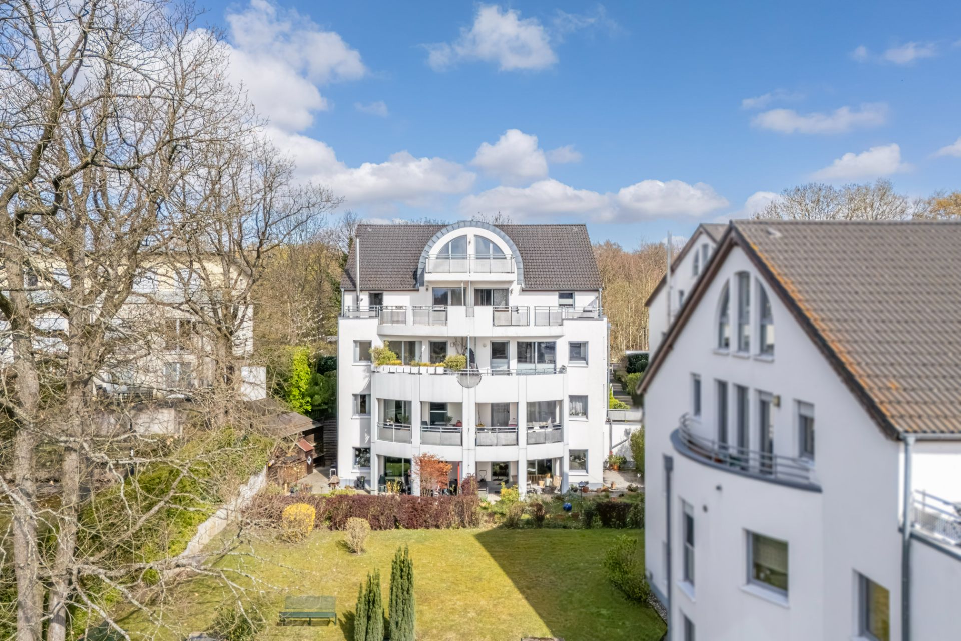 Modernes weißes Wohngebäude mit Balkonen und Garten im Vordergrund, Bäume und blauer Himmel.