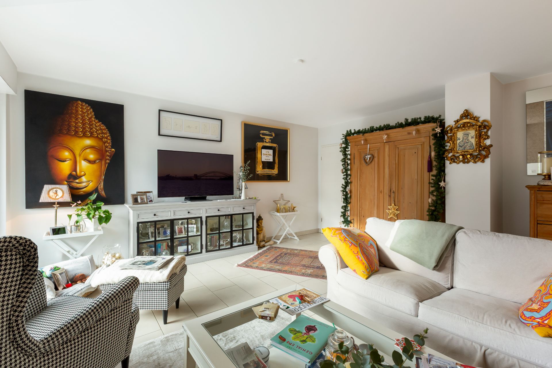 Helles Wohnzimmer mit Sofa, Sessel, Glastisch, TV, Gemälden und großem Holzschrank.