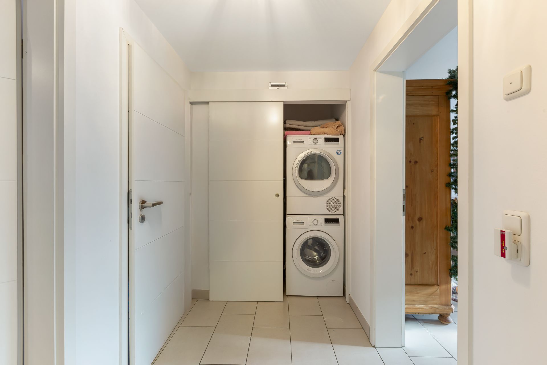Ein Flur mit Waschmaschine und Trockner in einem Wandschrank, daneben Türen und gefliester Boden.