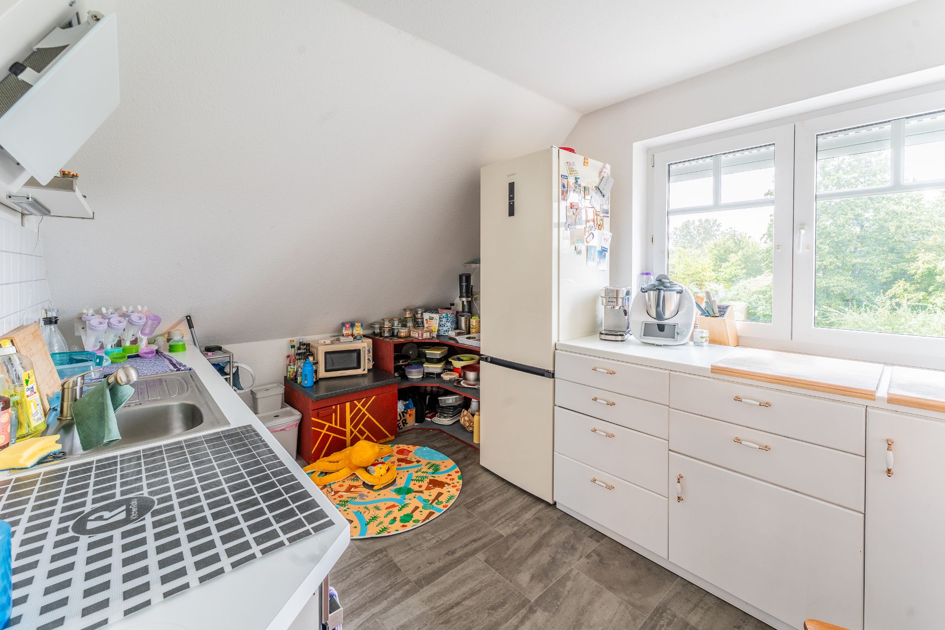 Helle Küche mit Spüle, weißem Kühlschrank, Regalen und großem Fenster mit Blick ins Grüne.