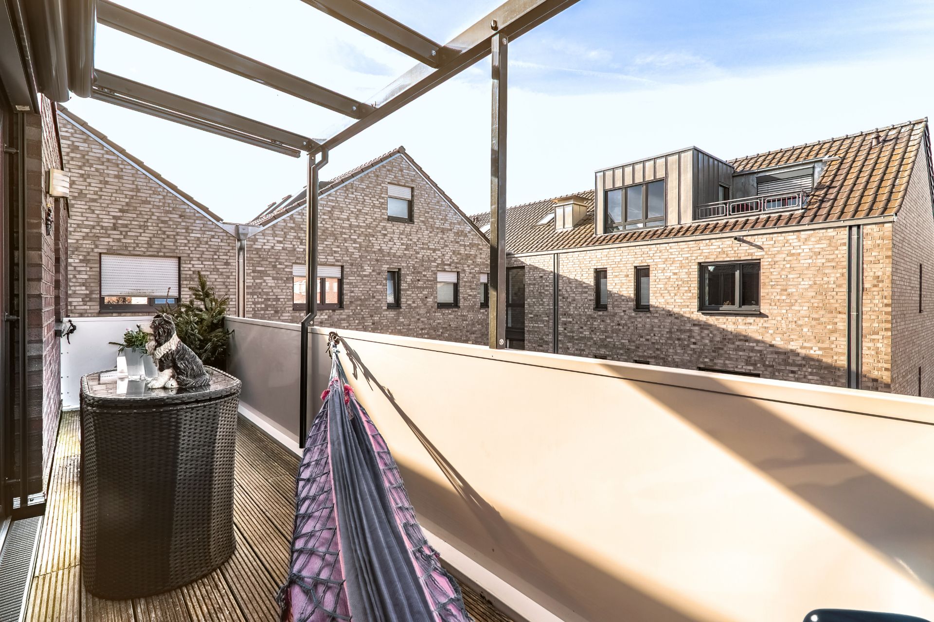 Balkon mit Holzboden, Tisch, Dekohund, Hängematte und Blick auf benachbarte Häuser.