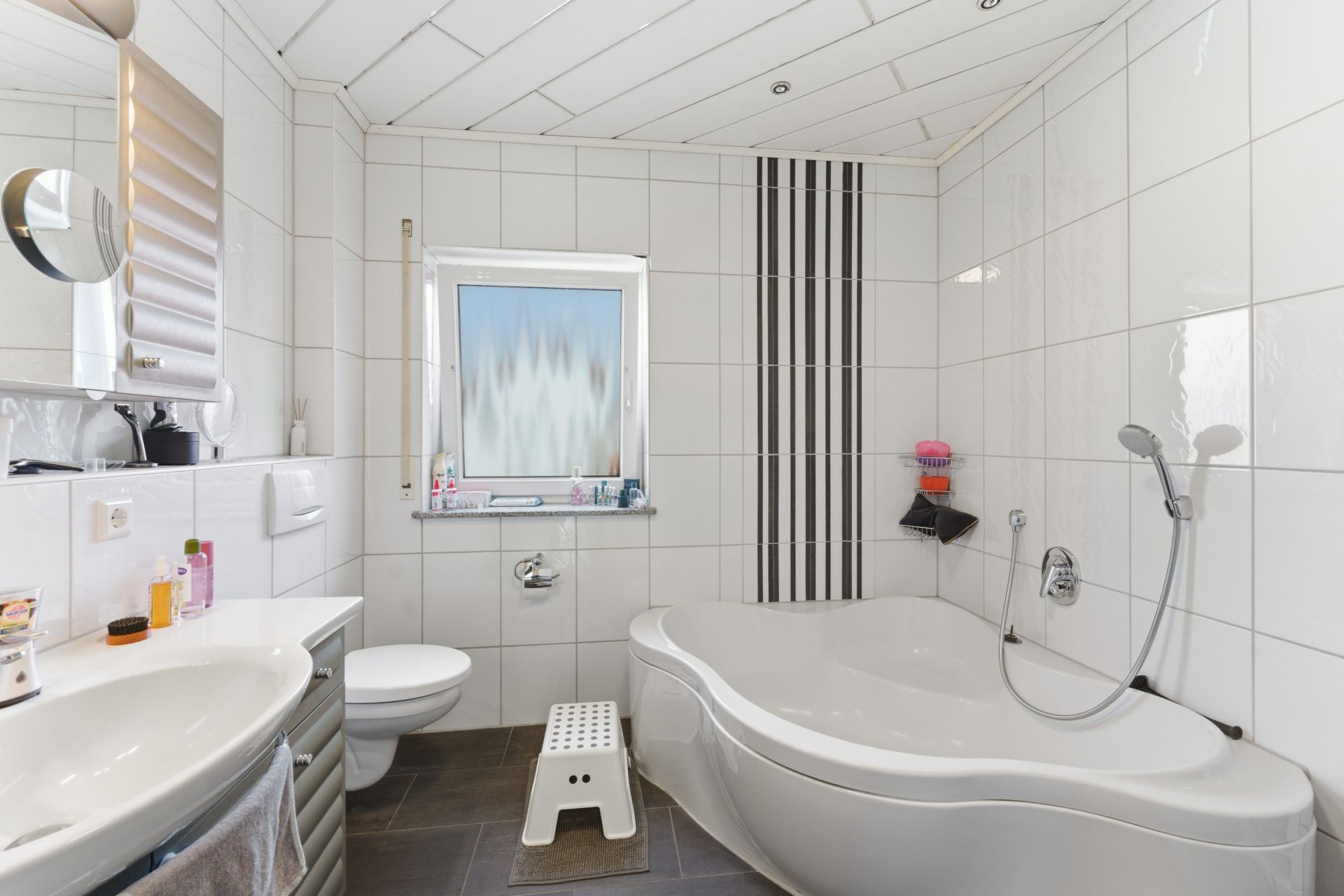 Modernes Badezimmer mit Eckbadewanne, Waschbecken, WC, Fenster und Hocker.