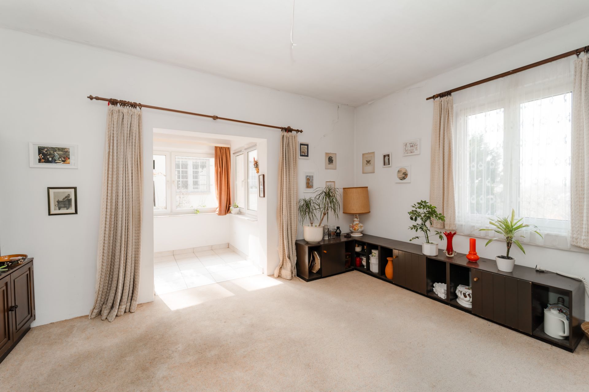 Helles Wohnzimmer mit Teppichboden, großen Fenstern, Vorhängen und Pflanzen auf einem Regal.
