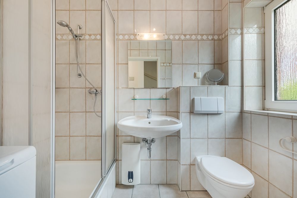 Helles Badezimmer mit Dusche, Waschbecken, WC und Fenster, alles mit hellen Fliesen ausgestattet.
