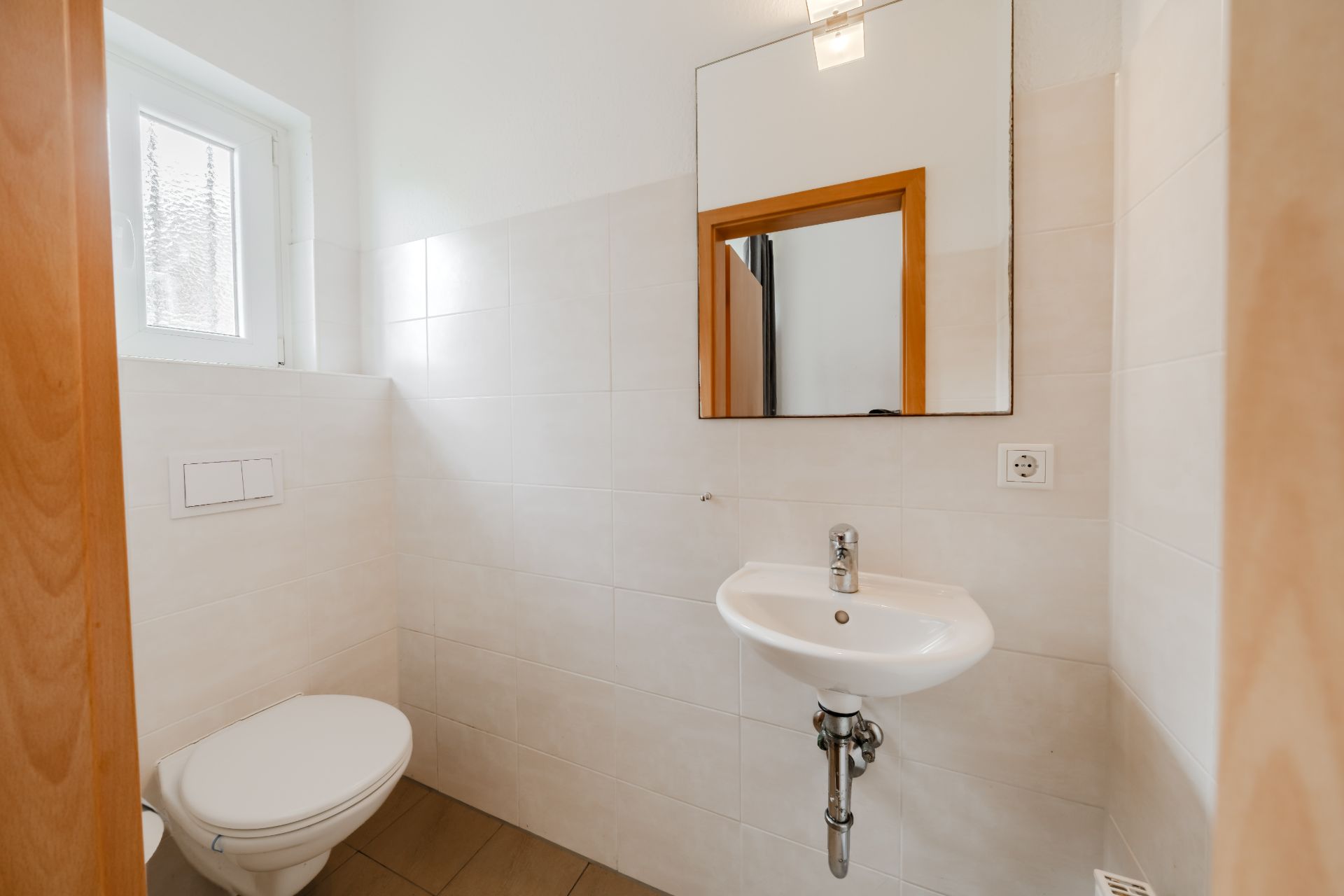 Kleines Badezimmer mit WC, Waschbecken und Spiegel, seitliches Fenster, helle Fliesen.