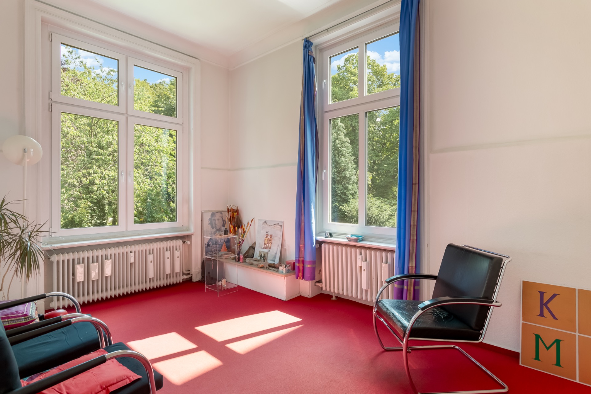 Helles Zimmer mit zwei großen Fenstern, roten Teppich, Stühlen und Pflanzen.