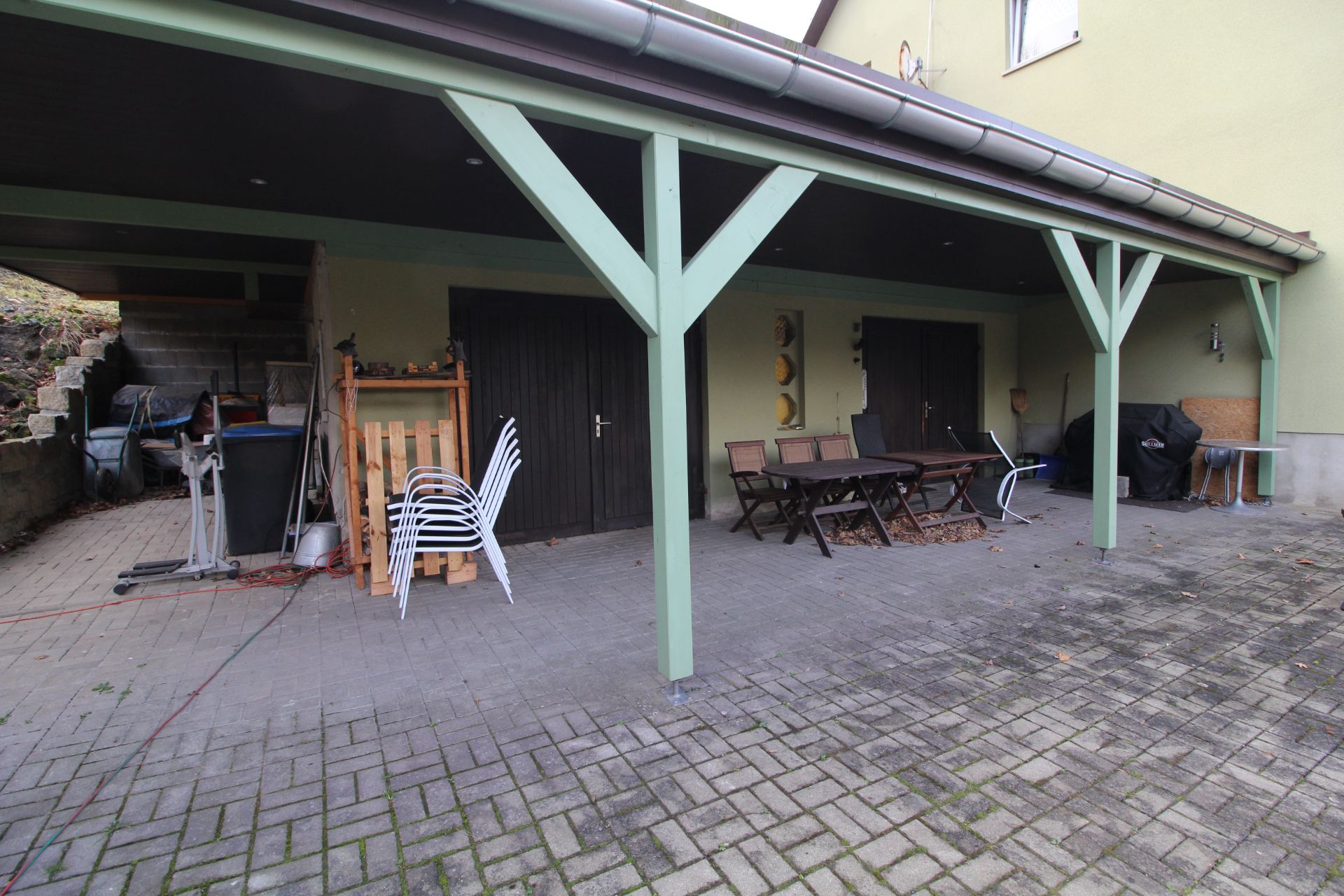 Überdachte Terrasse mit Gartentisch, Stühlen und zwei Türen, links gestapelte Gartenmöbel.