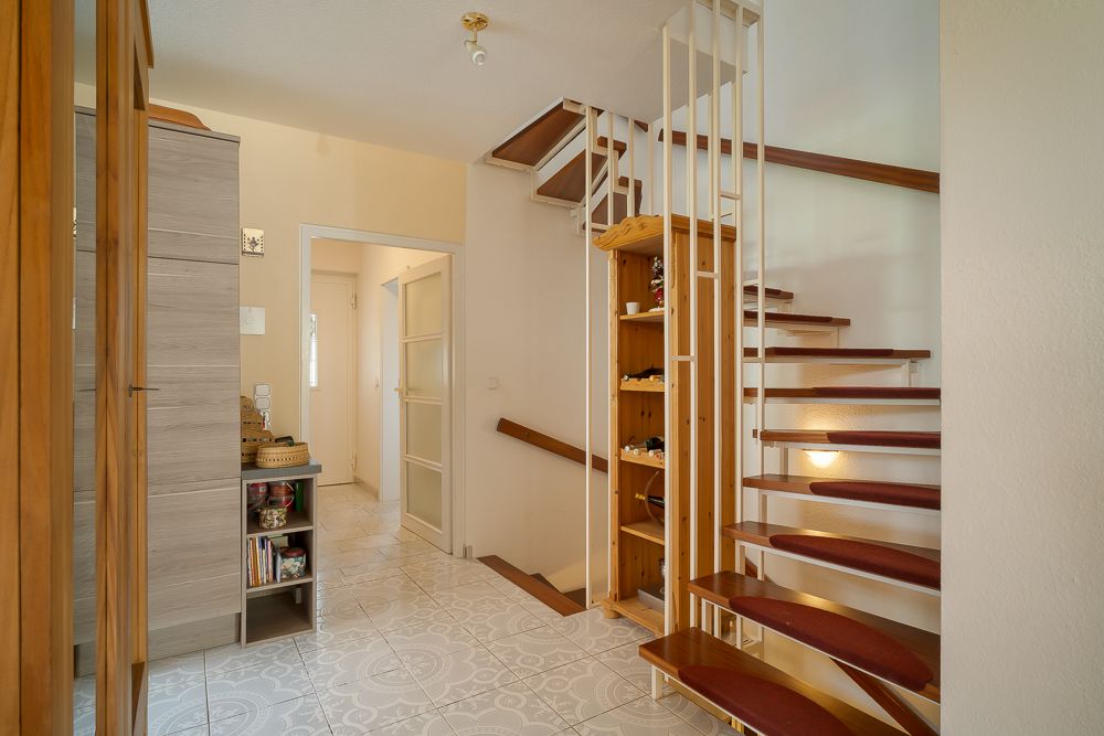 Heller Flur mit Holztreppe, Regal und Kleiderschrank, Fliesenboden, offene Tür zum Nebenraum.