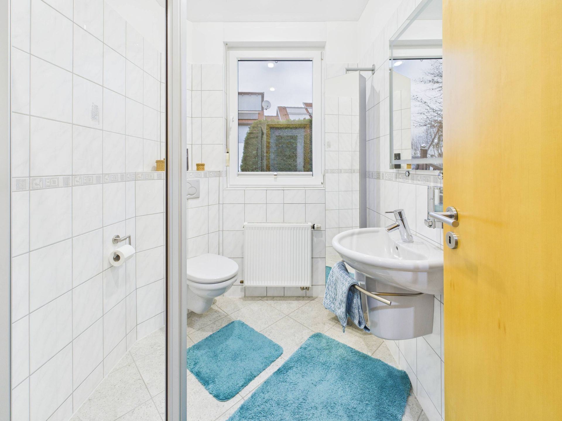 Helles Badezimmer mit weißem Fliesenspiegel, WC, Waschbecken, Fenster und zwei blauen Teppichen.