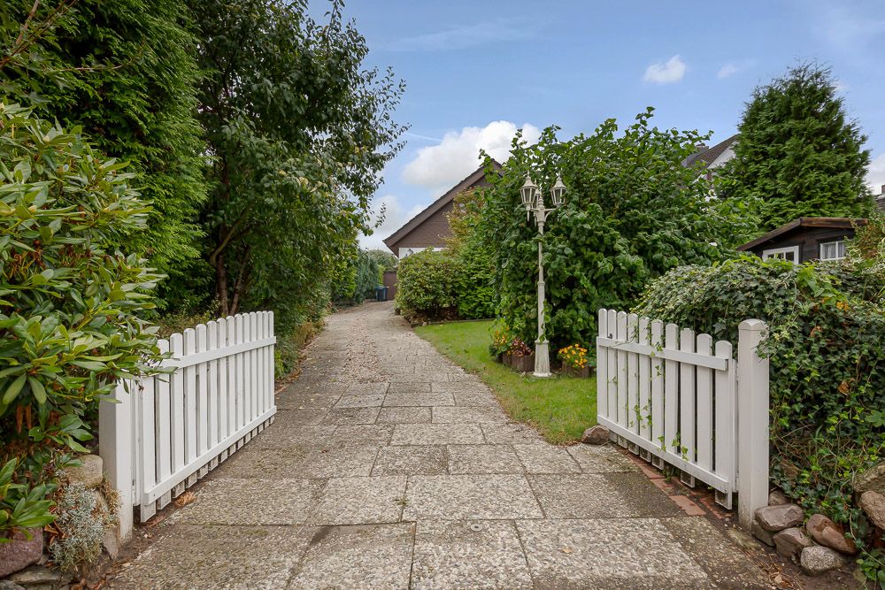 Geöffneter weißer Gartenzaun mit Zufahrtsweg, umgeben von Büschen und Bäumen, im Hintergrund ein Haus.
