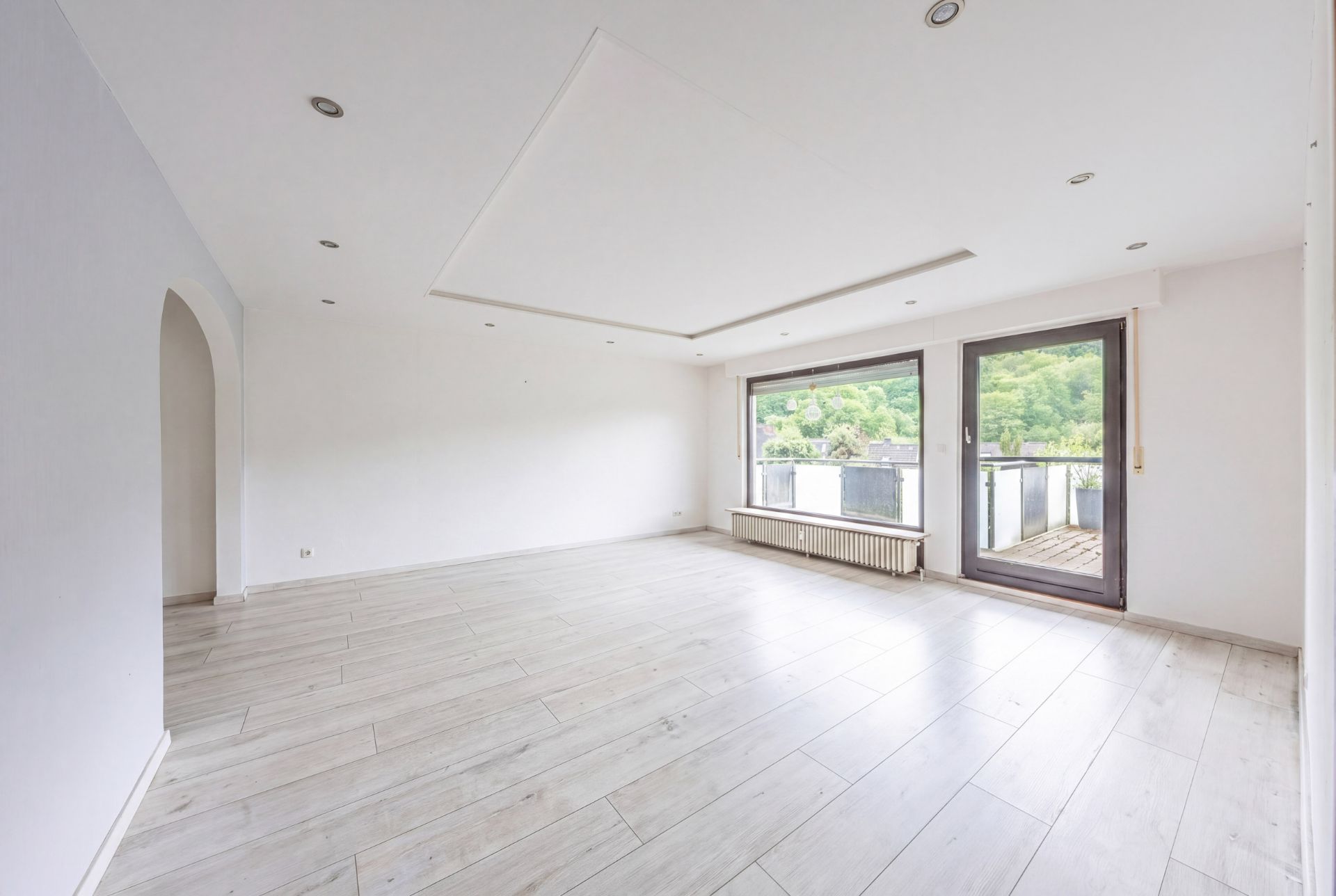 Helles, leeres Wohnzimmer mit großen Fenstern und Zugang zu einem Balkon.