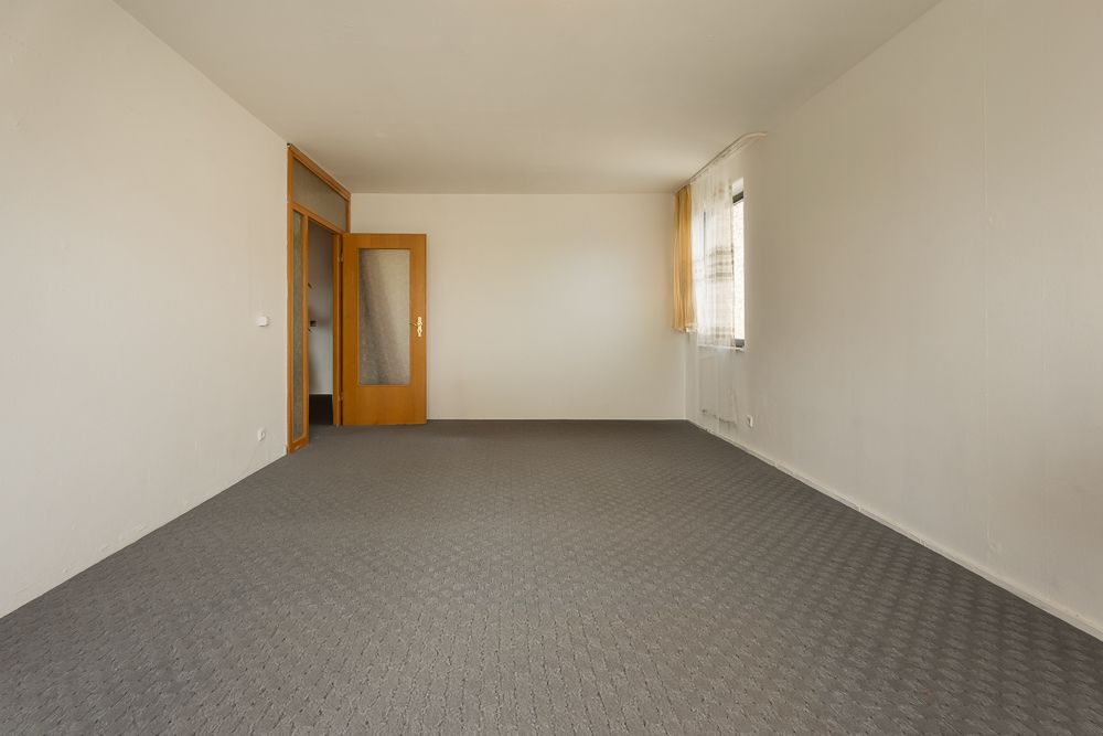 Leeres Zimmer mit grauem Teppich, weißen Wänden, Holztür und Fenster mit Vorhang.