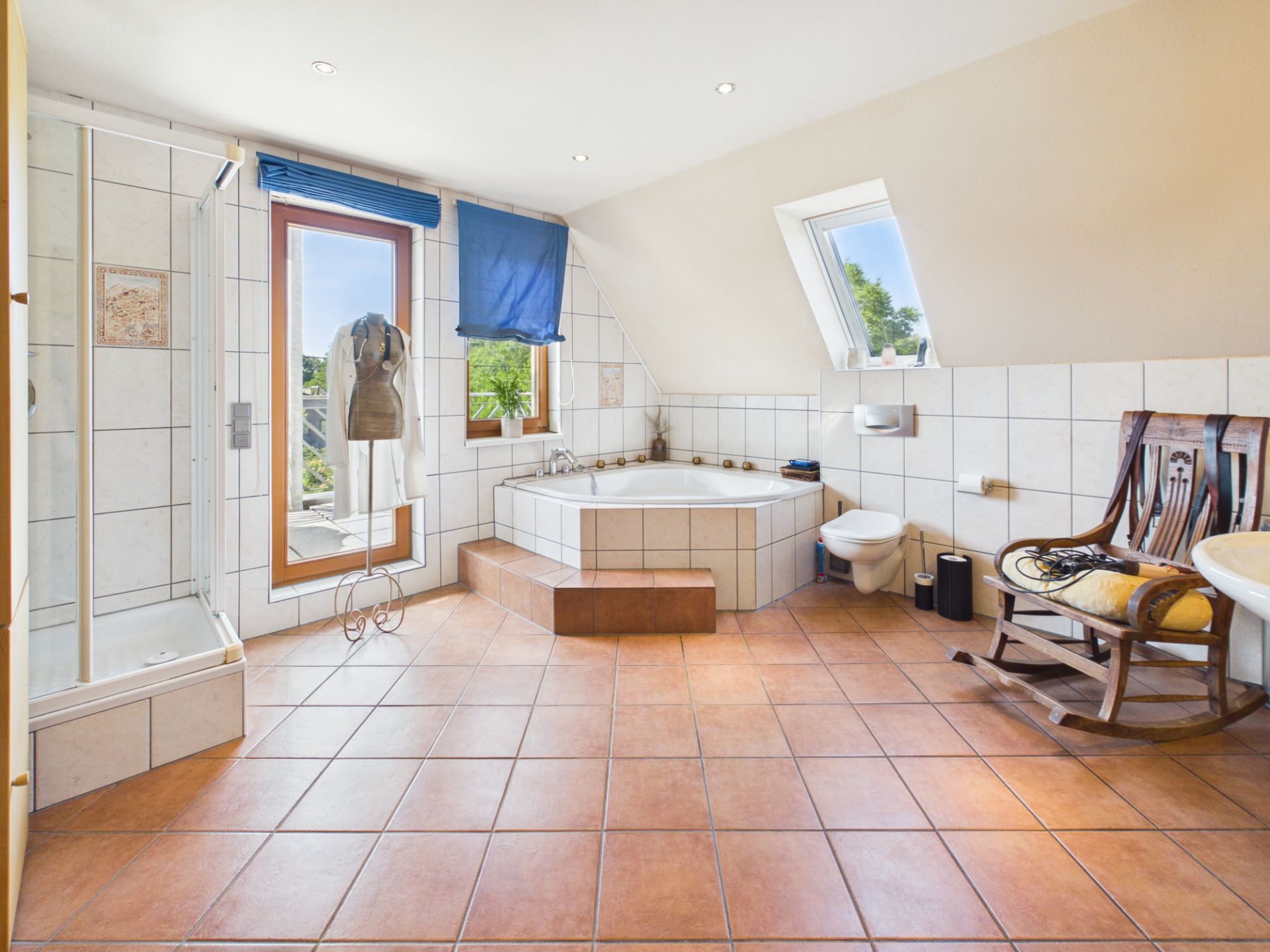 Großes Badezimmer mit Eckbadewanne, Dusche, WC, Holzstuhl und Fenster mit blauen Rollos.