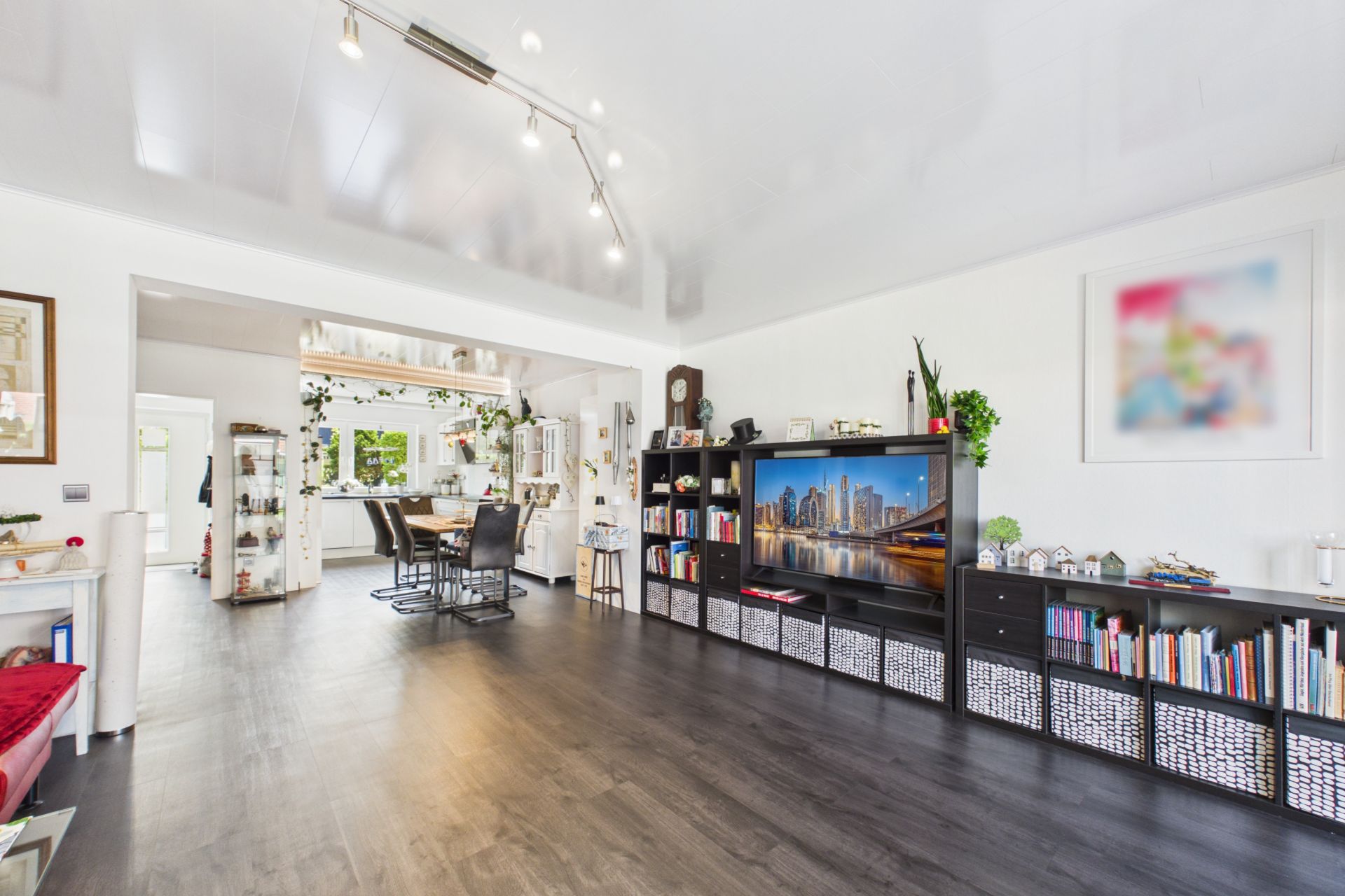 Helles Wohnzimmer mit dunklem Boden, Bücherregalen, großem Fernseher und offener Küche im Hintergrund.