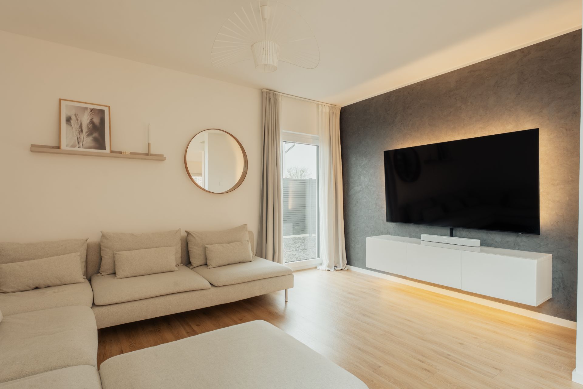 Modernes Wohnzimmer mit beigem Sofa, großem Fernseher, rundem Spiegel und Holzfußboden.