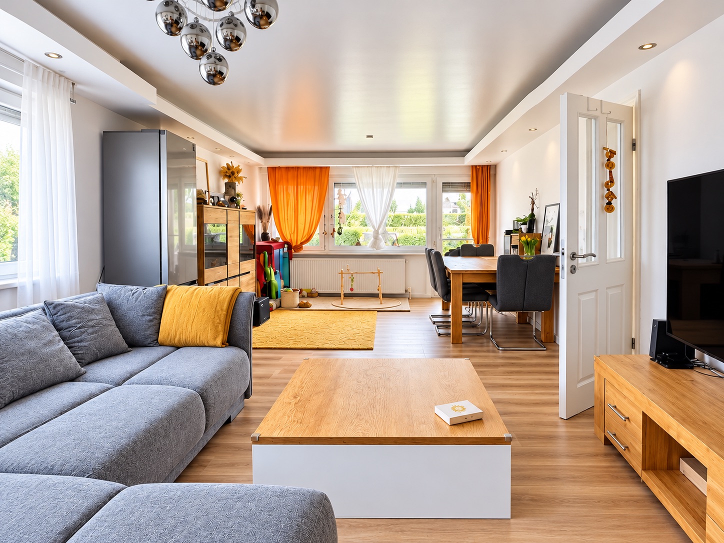 Helles Wohnzimmer mit grauem Sofa, Holzmöbeln, Esstisch und großen Fenstern mit orangefarbenen Vorhängen.