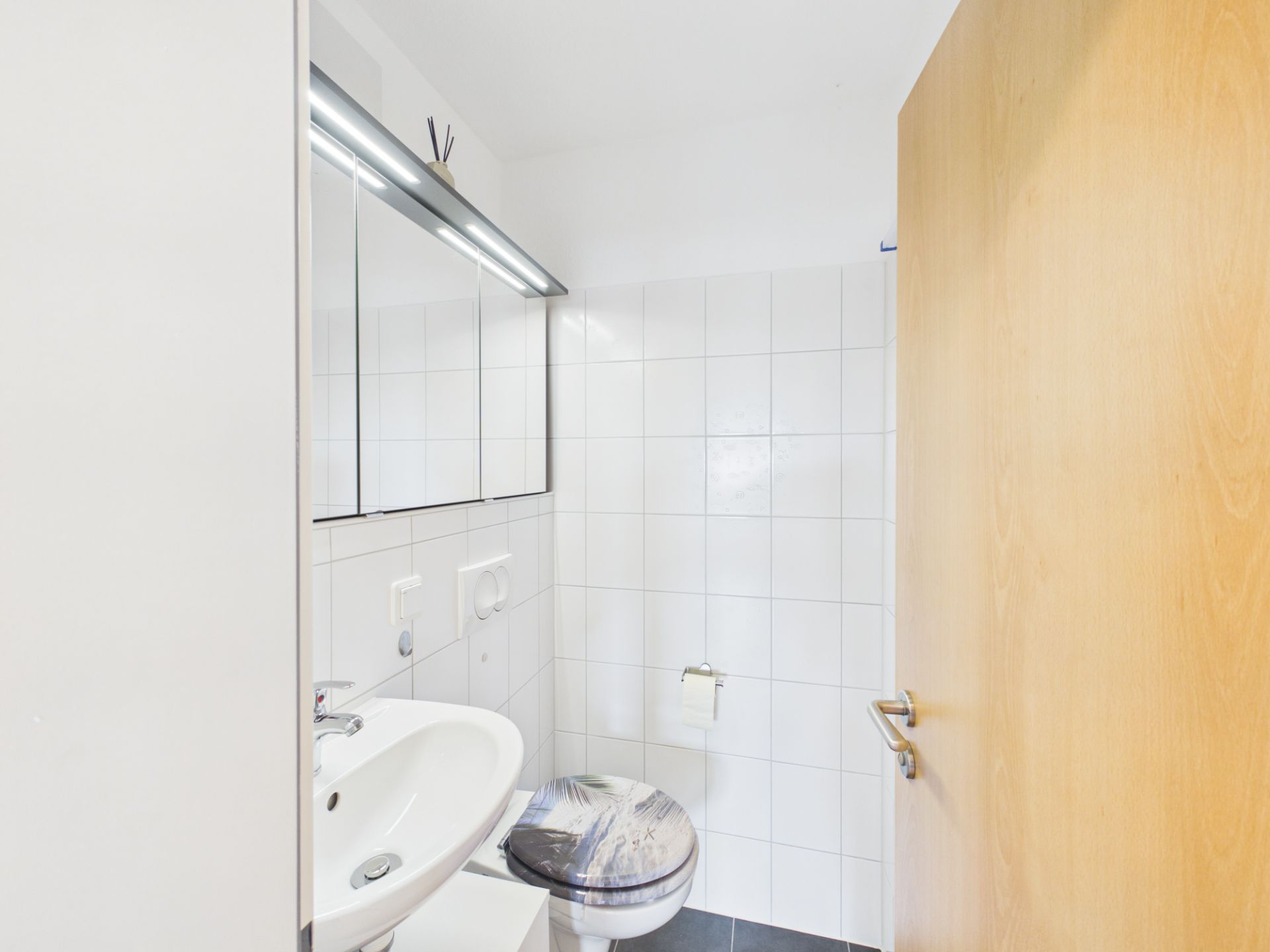 Kleines weiß gefliestes Badezimmer mit Waschbecken, Spiegel und Toilette.