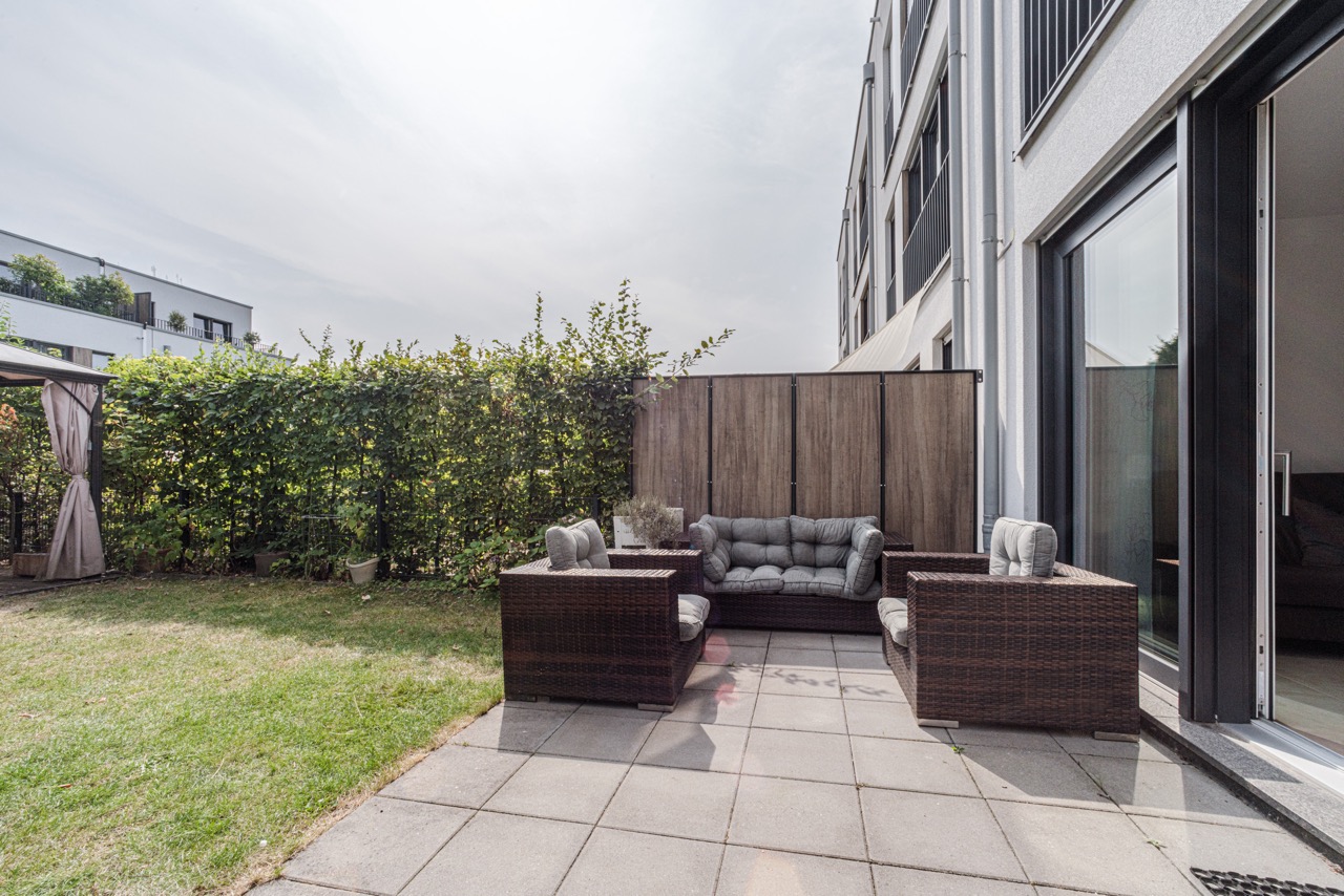 Terrasse mit Gartensesseln und Sofa, angrenzender Rasen und Sichtschutz, Wohnhaus im Hintergrund.