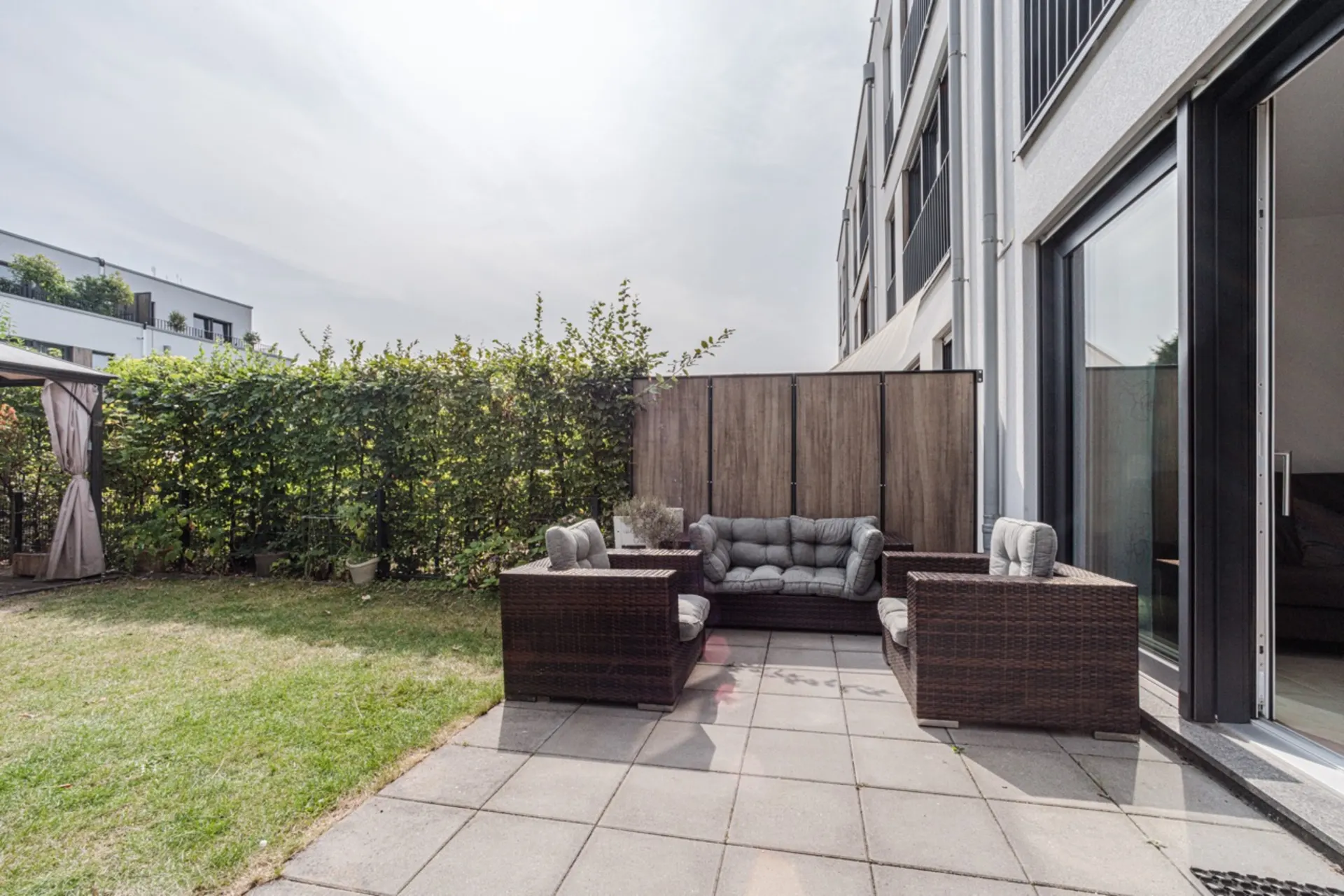 Terrasse mit Gartensesseln und Sofa, angrenzender Rasen und Sichtschutz, Wohnhaus im Hintergrund.