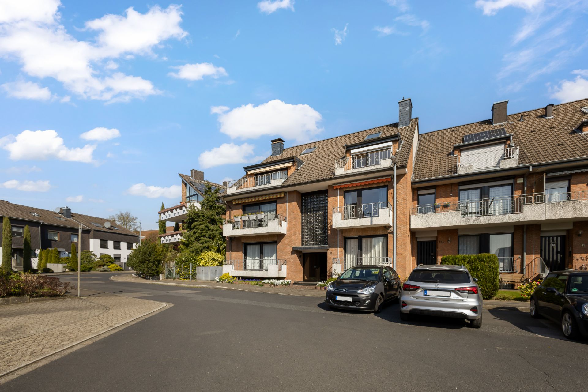 Mehrfamilienhaus mit Balkonen, geparkten Autos und blauem Himmel im Wohngebiet.