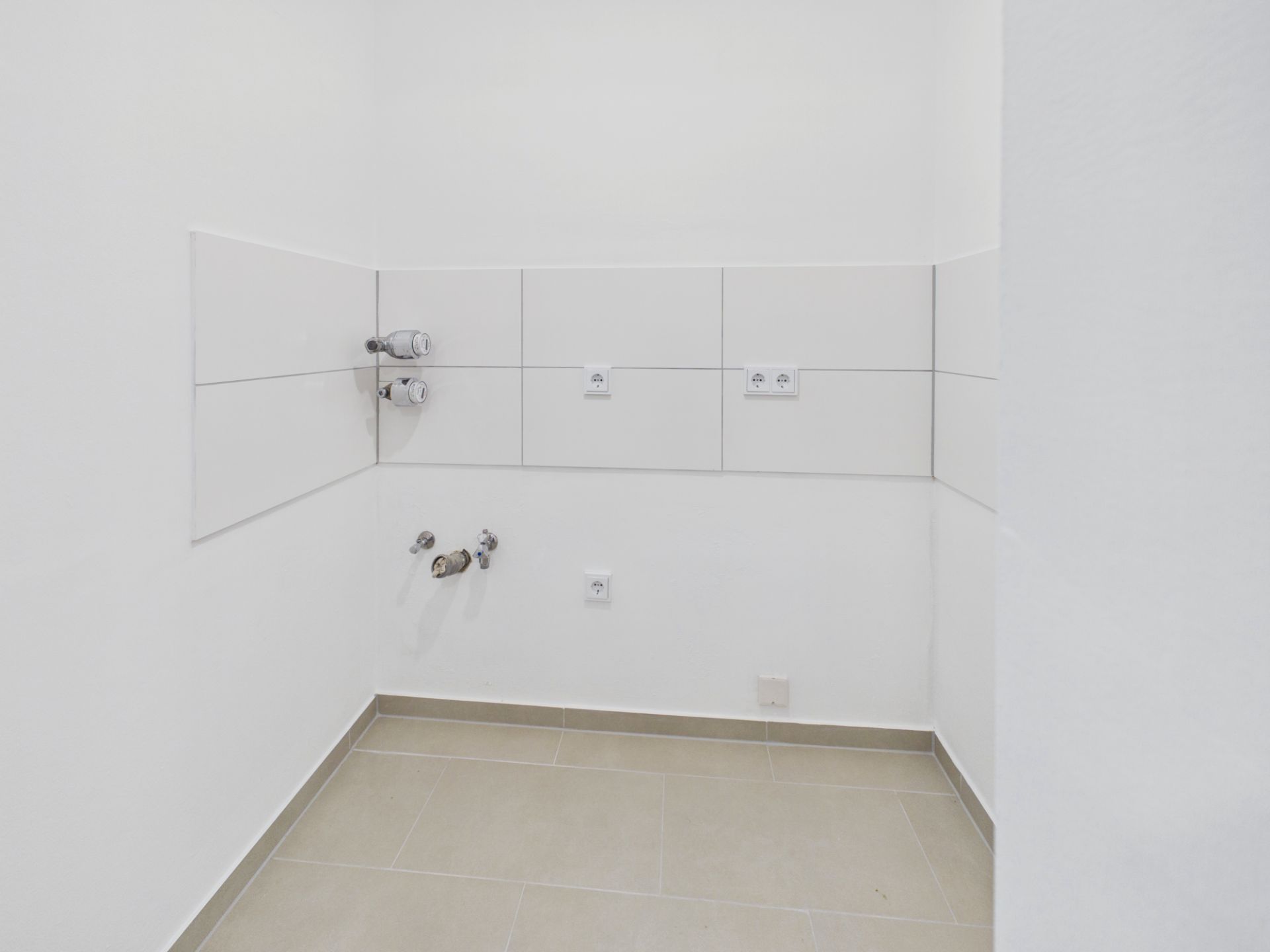Leerer Raum mit Fliesenboden, weißen Wänden und Anschlüssen für Wasser und Strom.