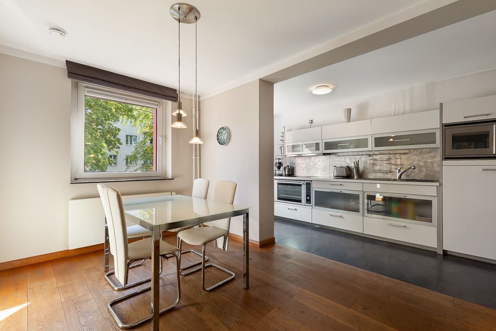 Moderne Küche mit Essbereich, Glastisch und vier Stühlen auf Holzboden, helles Fenster.