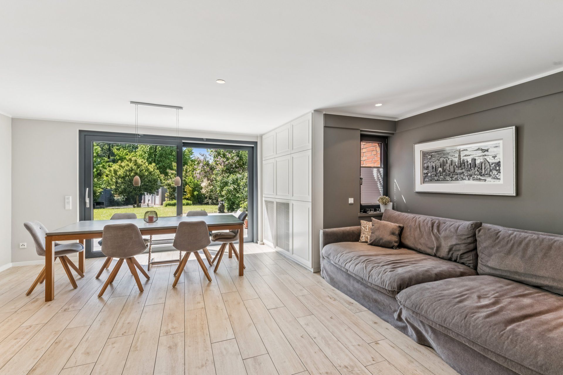 Modernes Wohnzimmer mit grauem Sofa, Essbereich und großem Fenster mit Blick auf den Garten.
