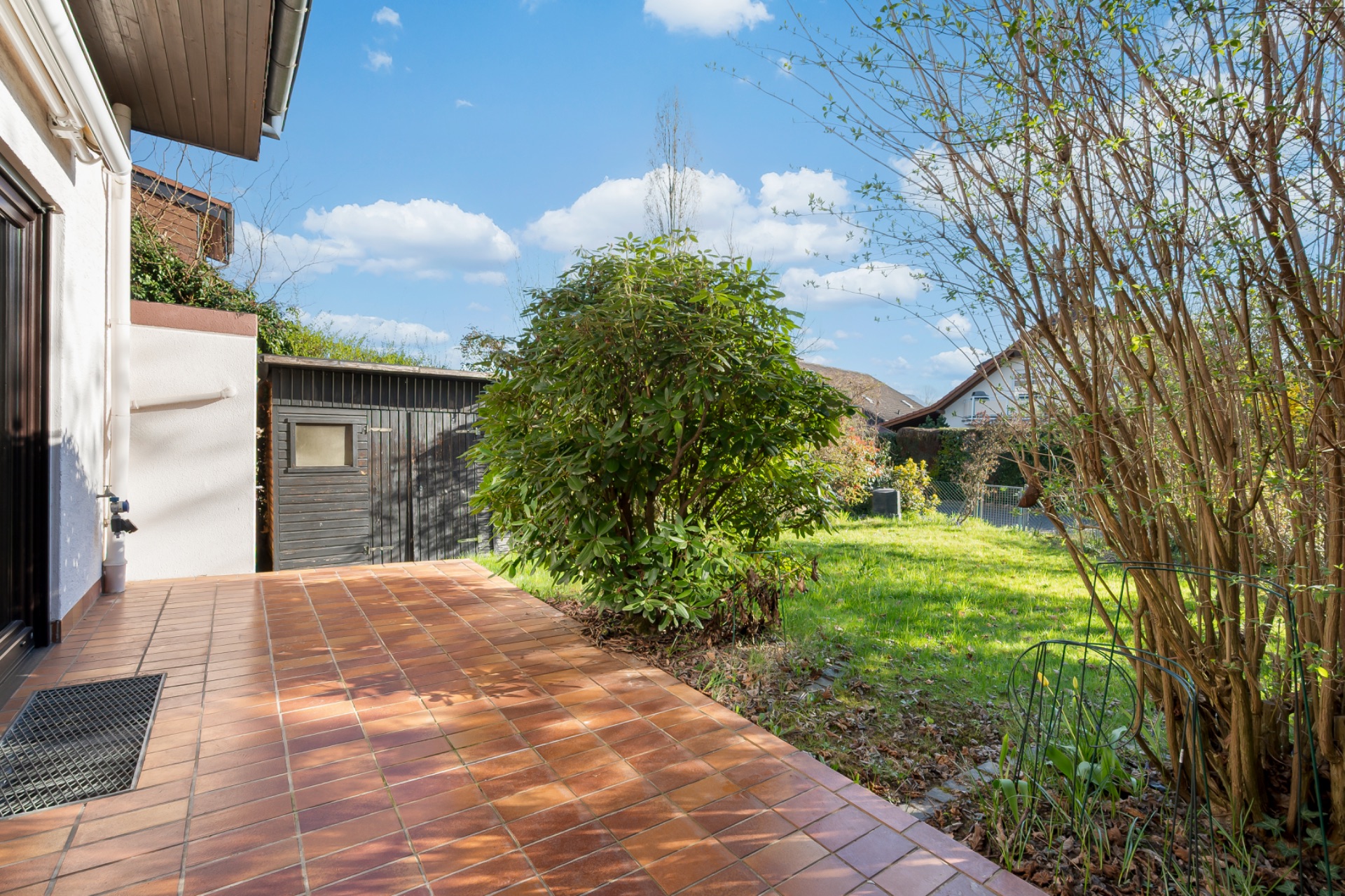 Geflieste Terrasse mit Blick auf Garten, Sträucher und ein Gartenhaus bei Tageslicht.