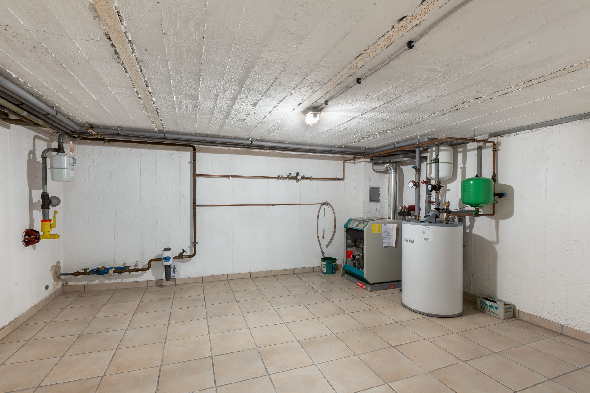 Kellerraum mit Fliesenboden, Heizungsanlage und einigen Rohrleitungen an der Wand.