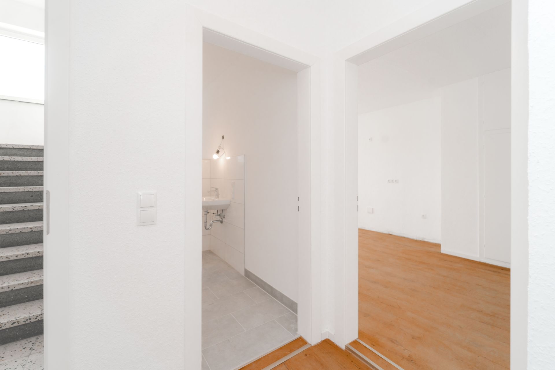 Flur mit Zugang zu einem Badezimmer und einem Raum mit Holzfußboden, Treppe links sichtbar.