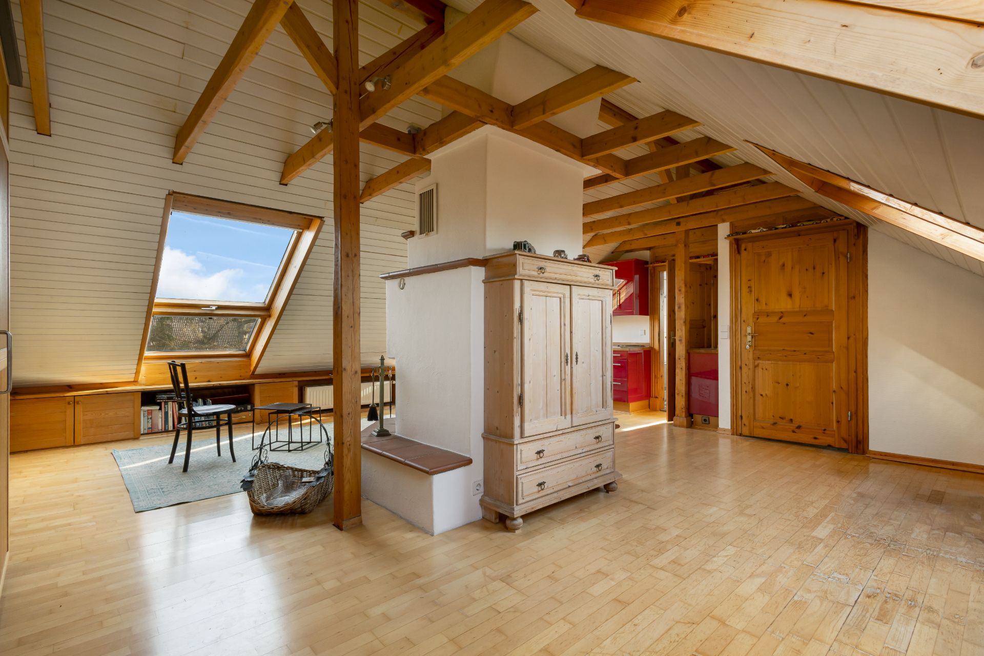 Helles Dachgeschosszimmer mit Holzbalken, Schreibtisch an einem großen Fenster und Holzschrank.