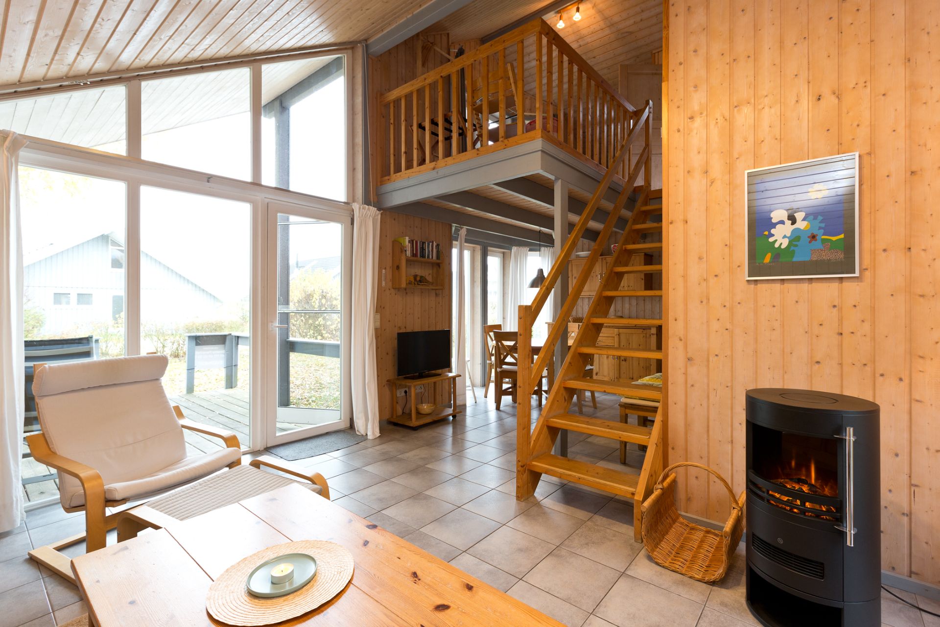 Helles Wohnzimmer mit Holztreppe, Kaminofen, Sessel, Tisch und großer Fensterfront.
