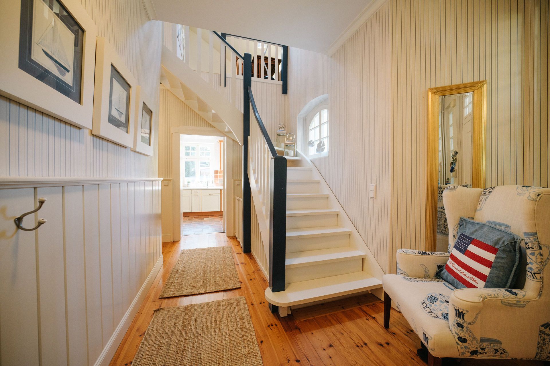 Heller Flur mit Treppe, Teppichen, Sessel und Wandbildern, im Hintergrund eine Küche.