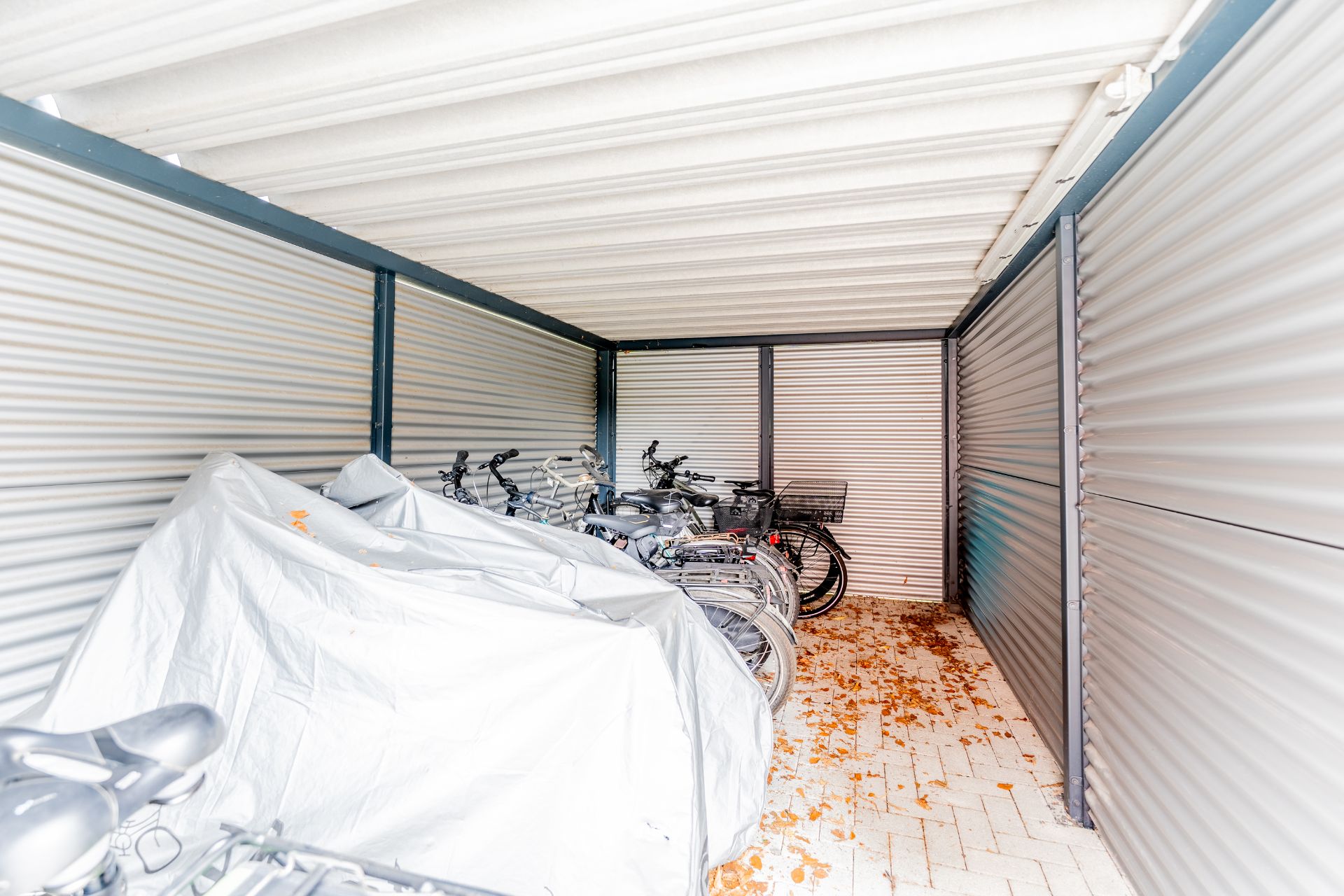Mehrere Fahrräder stehen in einem überdachten Fahrradunterstand mit Wellblechwänden.