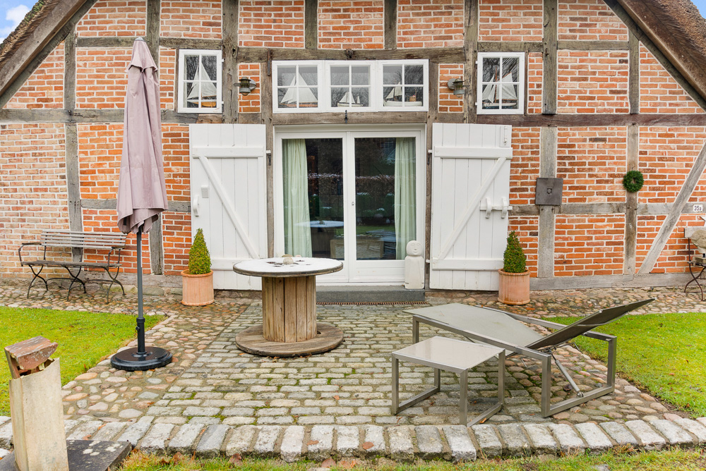 Gepflasterte Terrasse mit Tisch, Liege und Bank vor einem Fachwerkhaus mit weißen Fensterläden.