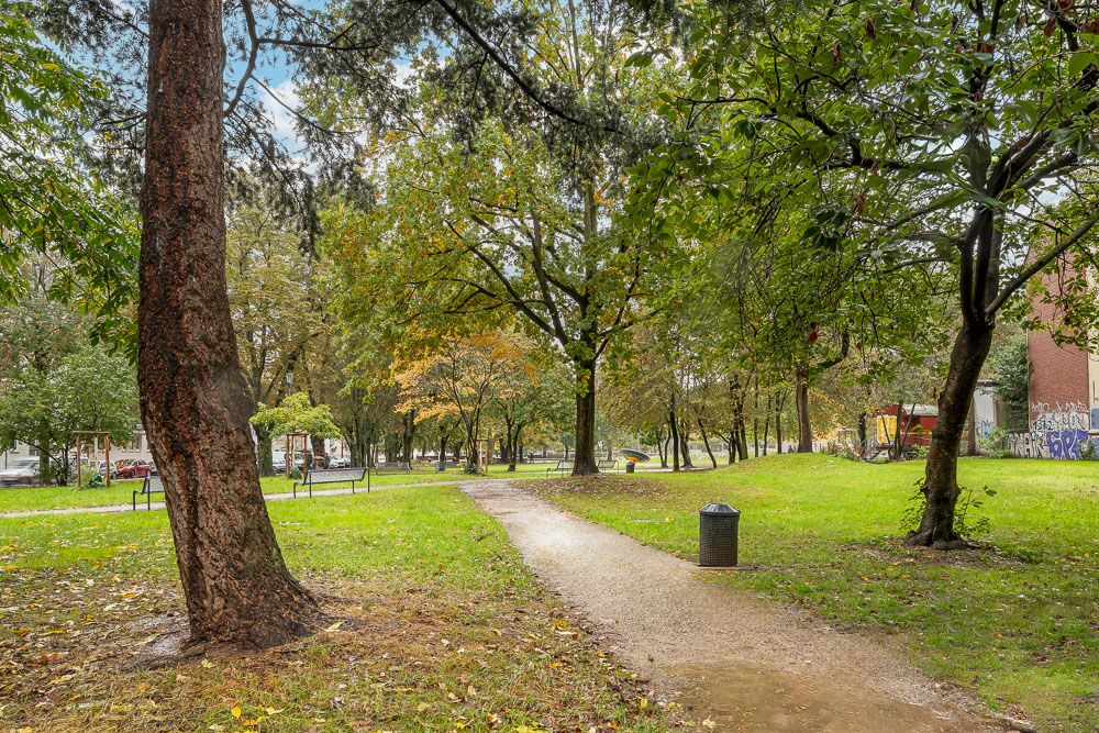 Schotterweg durch einen grünen Park mit Bäumen und Mülleimer.