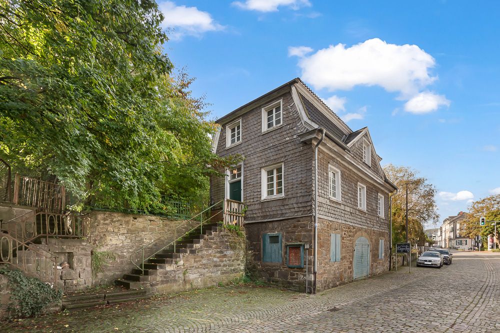 Zweistöckiges Haus mit grauer Holzfassade und Steintreppe, umgeben von Bäumen und Kopfsteinpflasterstraße.