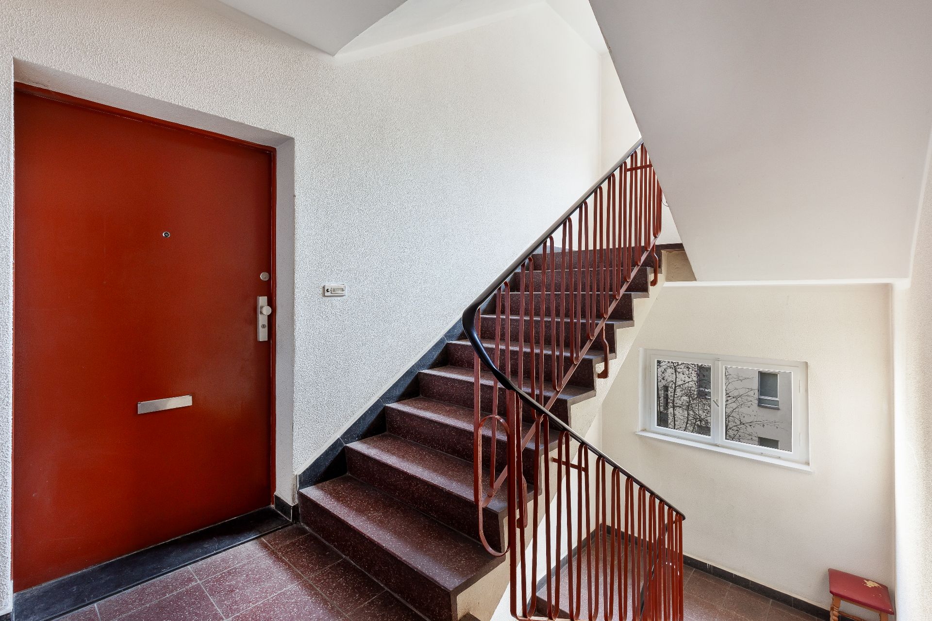 Treppenhaus mit roter Tür, rotbraunem Geländer und Fenster.