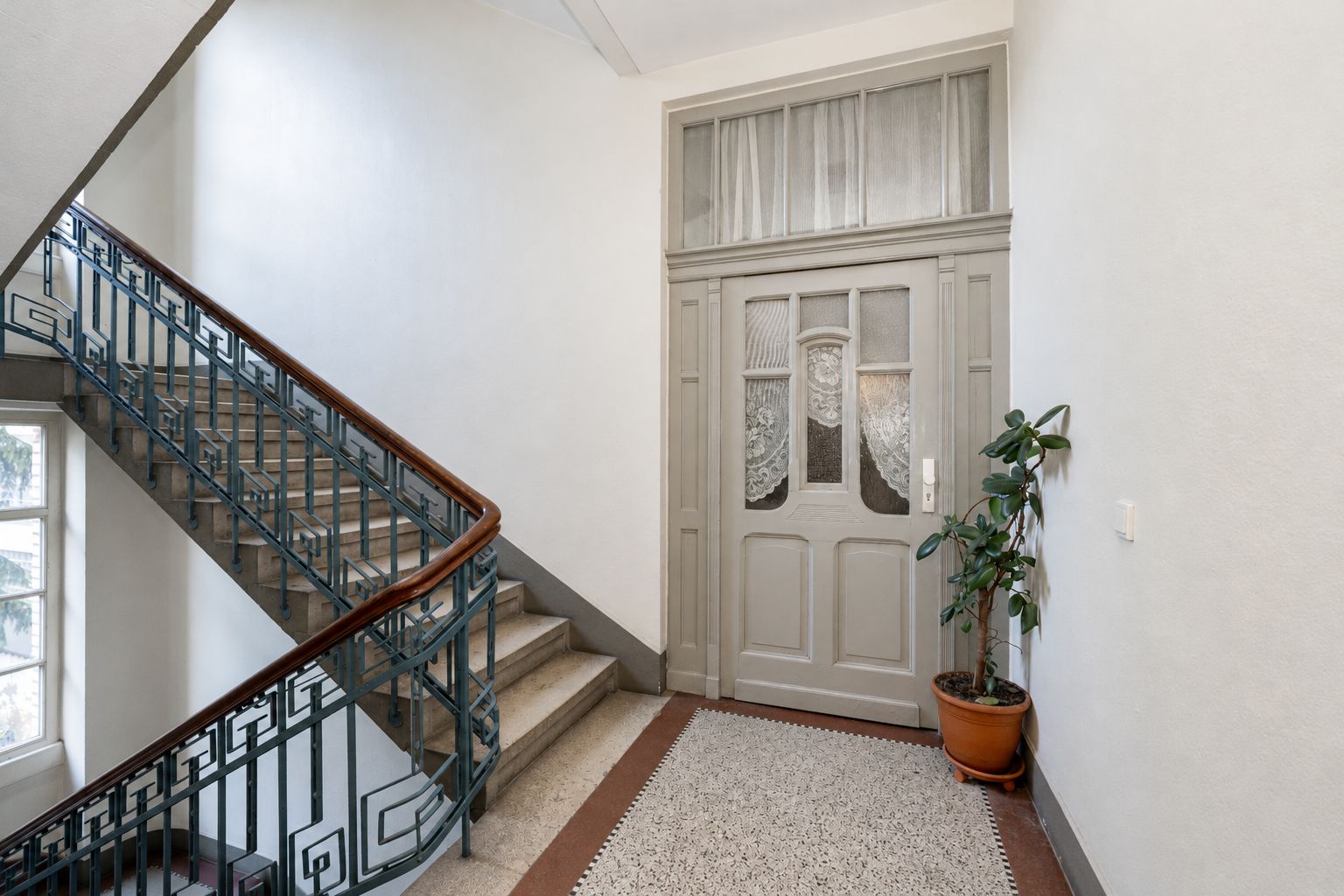 Treppenhaus mit Marmortreppe, Geländer, Eingangstür und einer Topfpflanze daneben.